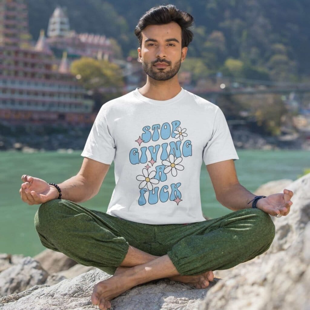 Man meditating on a rock wearing white t-shirt with a retro daisy and star design and the text 'Stop giving a fuck' printed on the front.