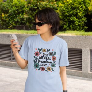 Woman wearing a light blue t-shirt with a multi color floral wreath design printed on the front. The words 'One mental breakdown later...' are printed in the center of the wreath.
