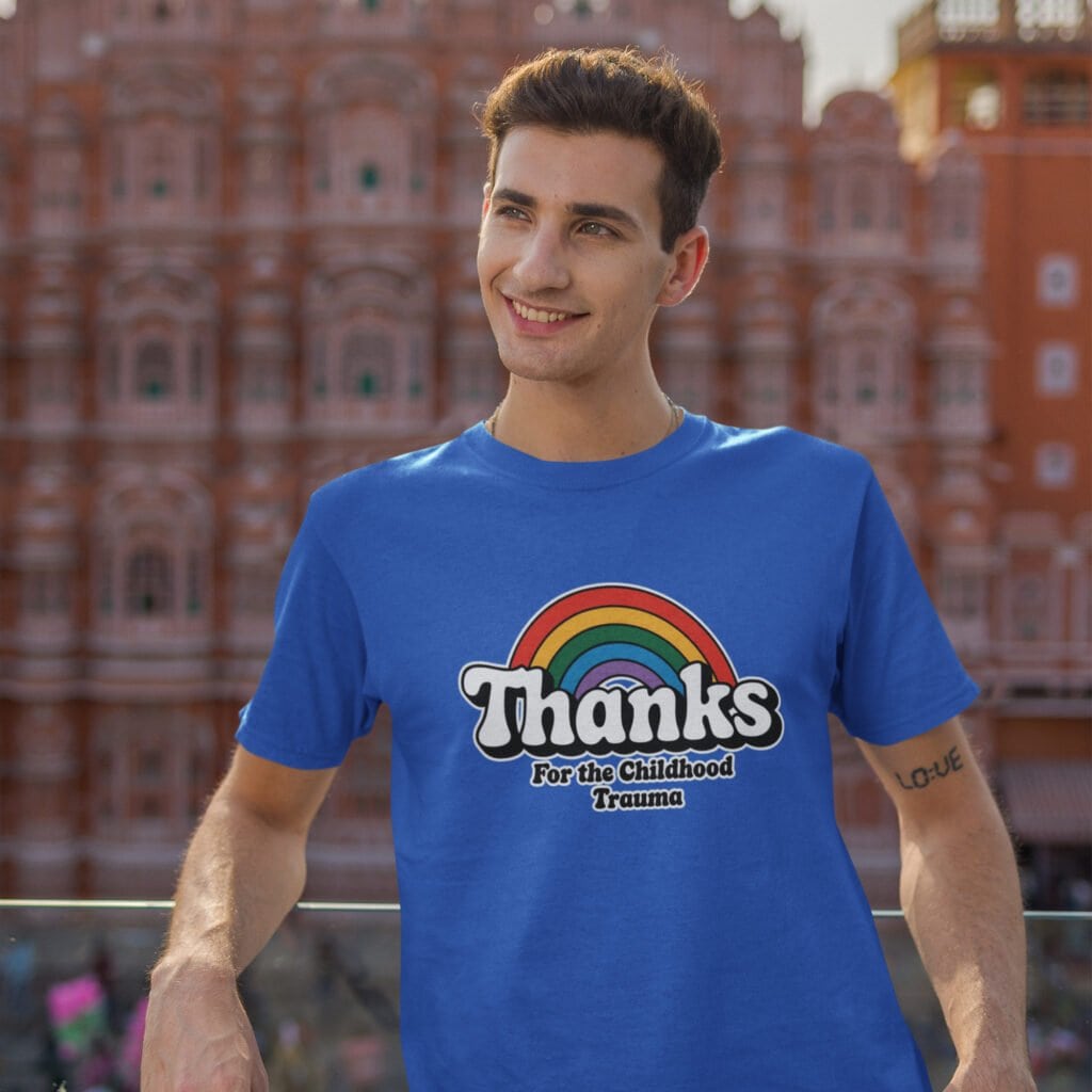 Man wearing a royal blue t-shirt with an image of a rainbow and the words 'Thanks for the childhood trauma' printed on the front. The word Thanks is large and the rest of the words are smaller under.