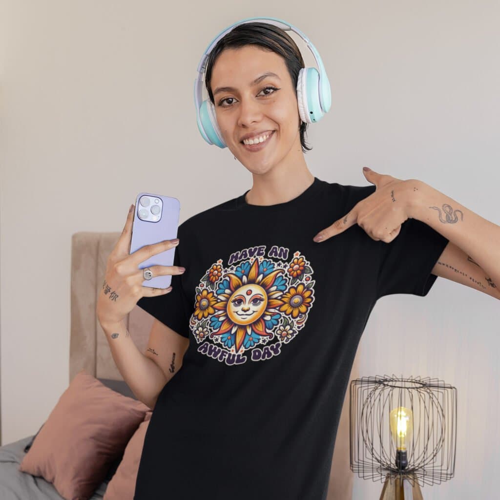 Woman wearing headphones and pointing at her shirt. She is wearing a black t-shirt with a sun graphic and the text 'Have an awful day' printed on the front.