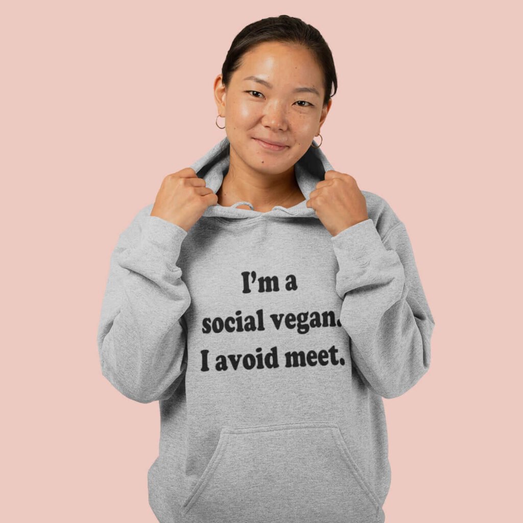 Woman wearing a light grey hoodie sweatshirt with the pun phrase 'I'm a social vegan. I avoid meet.' printed on the front.