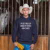 Man wearing a cowboy hat standing in a stable. He is wearing a navy blue hoodie sweatshirt with the funny phrase 'I'm so tired of being my wife's arm candy' printed on the front.