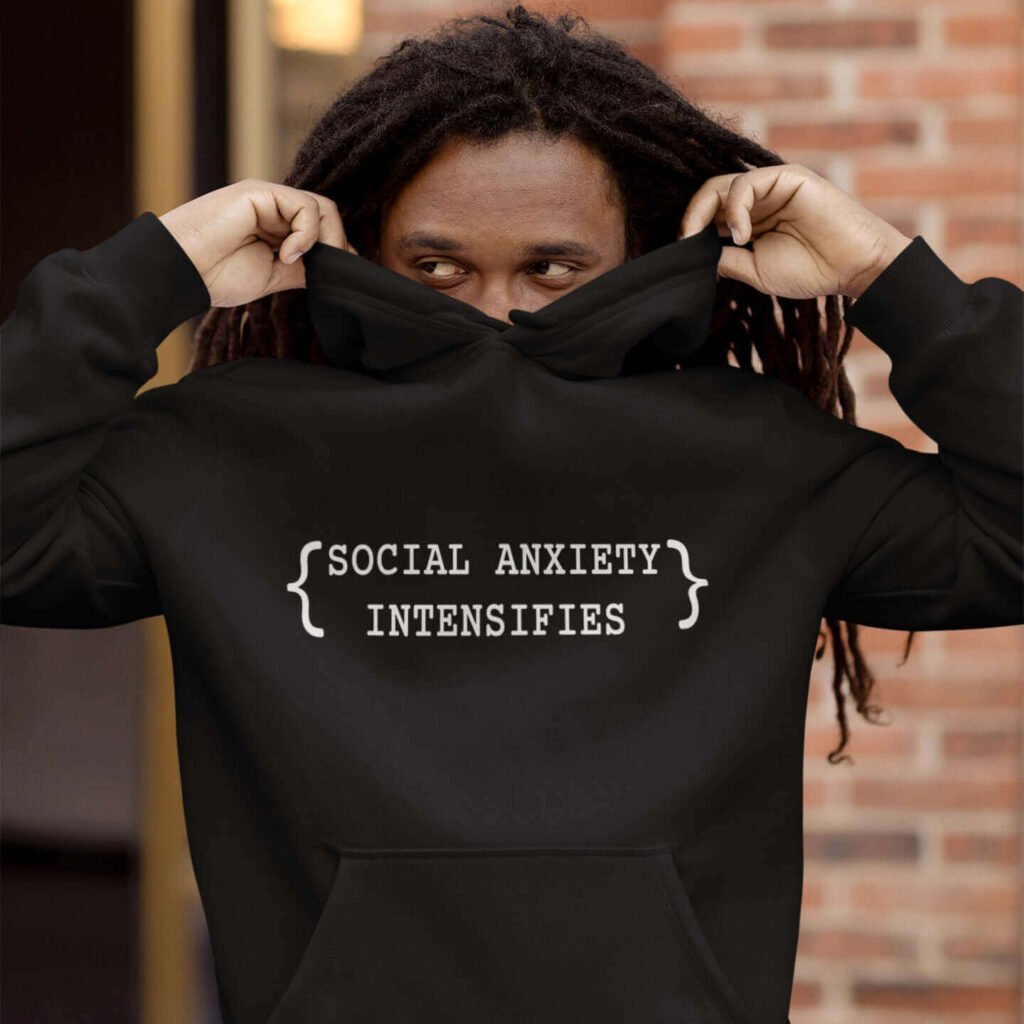 Man hiding his face in a hoodie sweatshirt. The hoodie has the text 'Social anxiety intensifies' printed on the front.