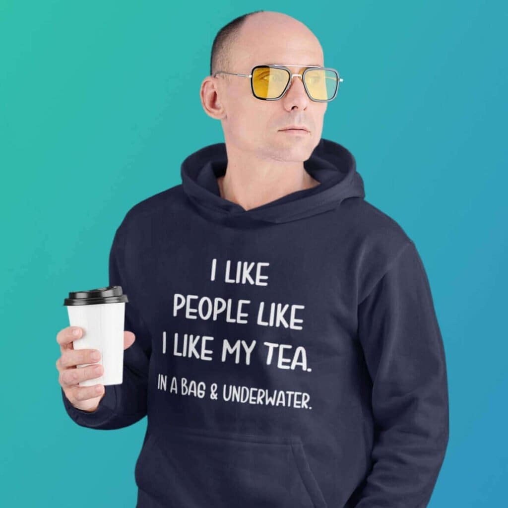 Serious looking man holding cup of tea. He is wearing a navy blue hoodie sweatshirt with the text 'I like people like I like my tea. In a bag & underwater.' printed on the front.