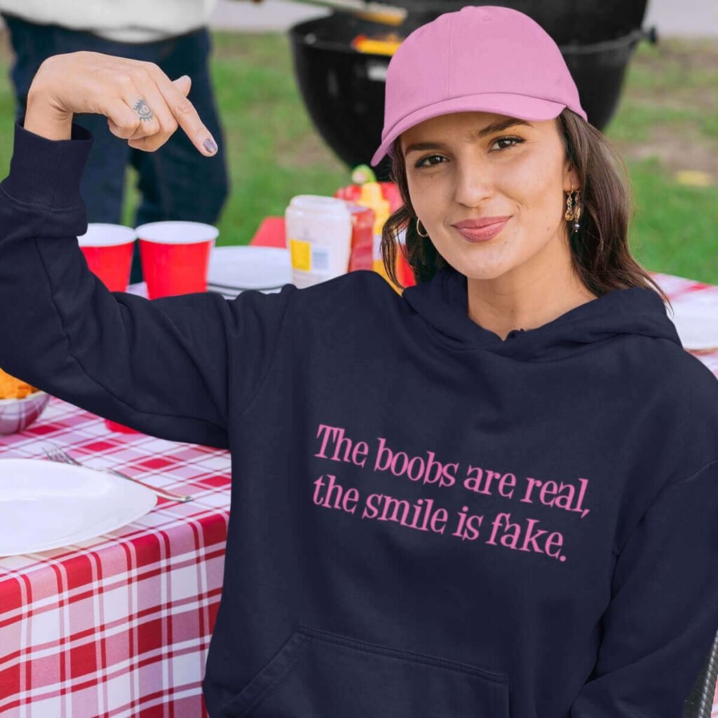 Woman wearing a navy blue hoodie sweatshirt with the text 'The boobs are real, the smile is fake.' printed in pink on the front.