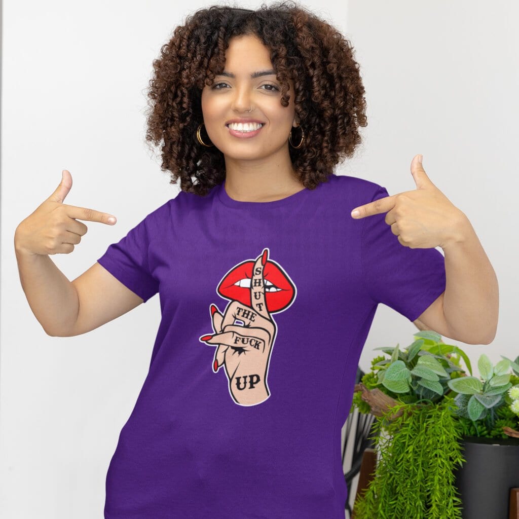 Woman wearing a purple t-shirt with an image of a hand over lips making the shh gesture. The words 'Shut the fuck up' are printed on the hand. The graphics are printed on the front.