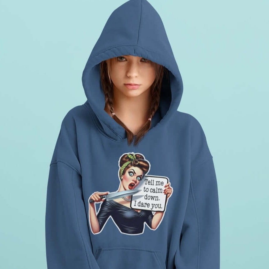 Woman wearing an indigo blue hooded sweatshirt with the hood on. The sweatshirt has a graphic of an angry looking retro woman holding a knife and a sign. The sign says 'Tell me to calm down. I dare you.' The graphic is printed on the front of the hoodie.