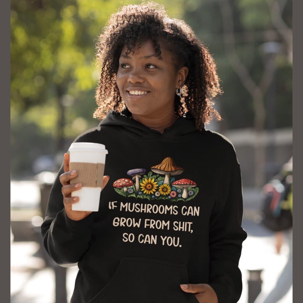 Smiling woman wearing a black hoodie sweatshirt with images of mushrooms and the text 'If mushrooms can grow from shit so can you' printed on the front.