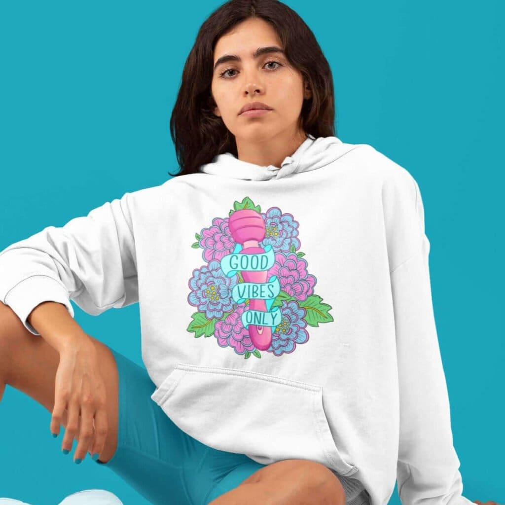 Woman wearing white hoodie sweatshirt with graphic design that has the words Good vibes only layered over a pink wand vibrator with flowers around. The graphic design is printed on the front of the hoodie.