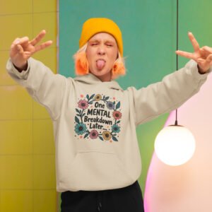 Woman wearing a sand color hoodie sweatshirt with a multi color floral wreath design printed on the front. The words 'One mental breakdown later...' are printed in the center of the wreath.