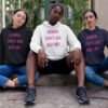 Three fierce looking women sitting next to each other. They are all wearing a hoodie sweatshirt with the text 'Women don't owe you shit' printed on the front in pink.
