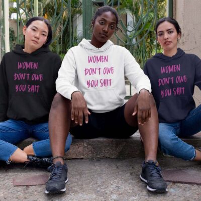 Three fierce looking women sitting next to each other. They are all wearing a hoodie sweatshirt with the text 'Women don't owe you shit' printed on the front in pink.