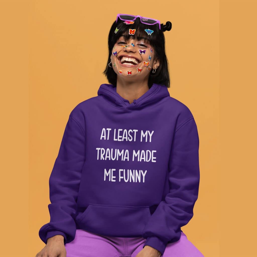 Woman wearing a Purple hoodie sweatshirt with the text 'At least my trauma made me funny' printed on the front.