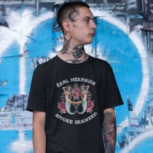 Tattooed man wearing a black t-shirt with image of 2 mermaids and the text 'Real mermaids smoke seaweed' printed on the front.