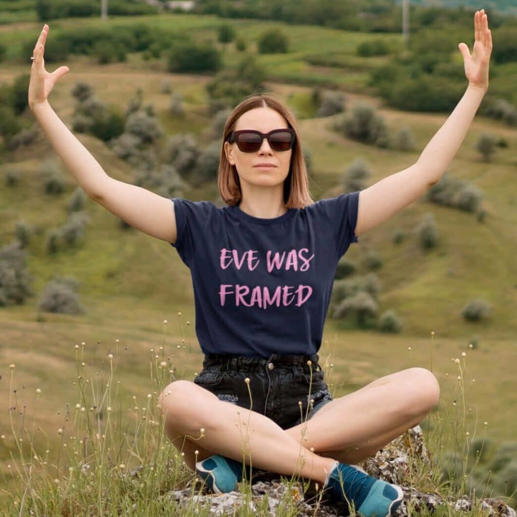 Woman sitting on a hill wearing a navy blue t-shirt with the text 'Eve was framed' printed on the front in pink.
