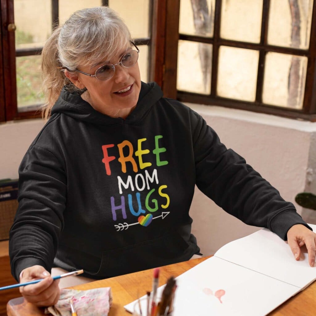 Older woman wearing black hoodie sweatshirt with the text 'Free Mom hugs' in rainbow lettering printed on the front.
