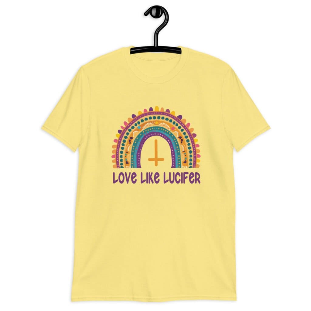Cornsilk yellow t-shirt with an image of a boho style rainbow. There is an upside down cross in the center arch of the rainbow and the text 'Love like Lucifer' printed under. The graphics are printed on the front.