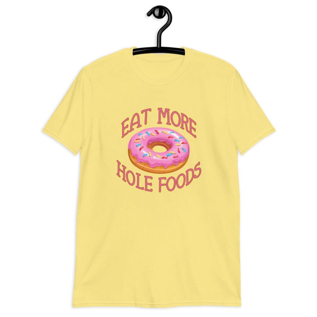 Cornsilk yellow t-shirt that has an image of a donut with pink icing and sprinkles and the text 'Eat more hole foods' printed on the front.