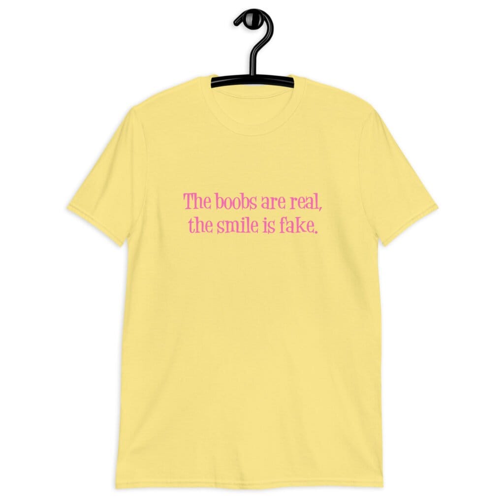Cornsilk yellow t-shirt with the text 'The boobs are real, the smile is fake.' printed in pink on the front.