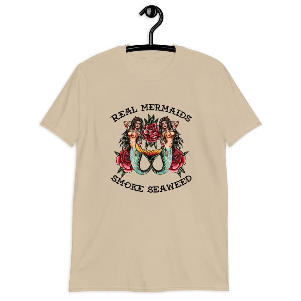 Sand color t-shirt with image of 2 mermaids and the text 'Real mermaids smoke seaweed' printed on the front.