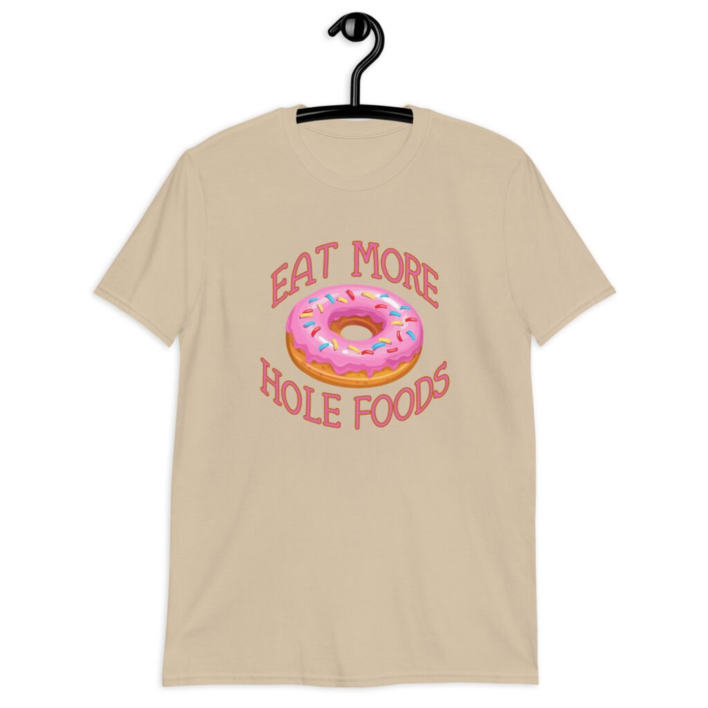 Sand color t-shirt that has an image of a donut with pink icing and sprinkles and the text 'Eat more hole foods' printed on the front.