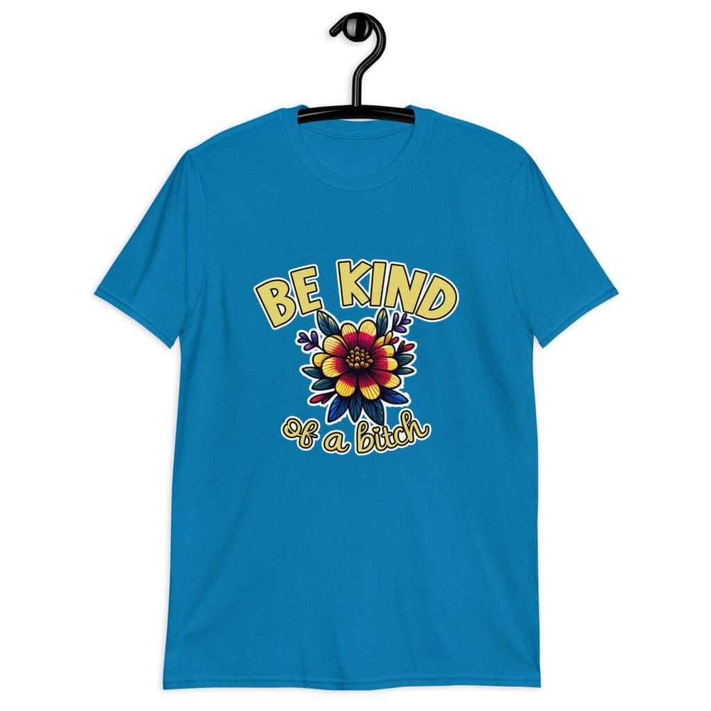 Sapphire bllue t-shirt with an image of a flower and the words Be kind above the flower in yellow bold block font. The words Of a bitch are smaller in script font under the flower. The design is printed on the front of the shirt.