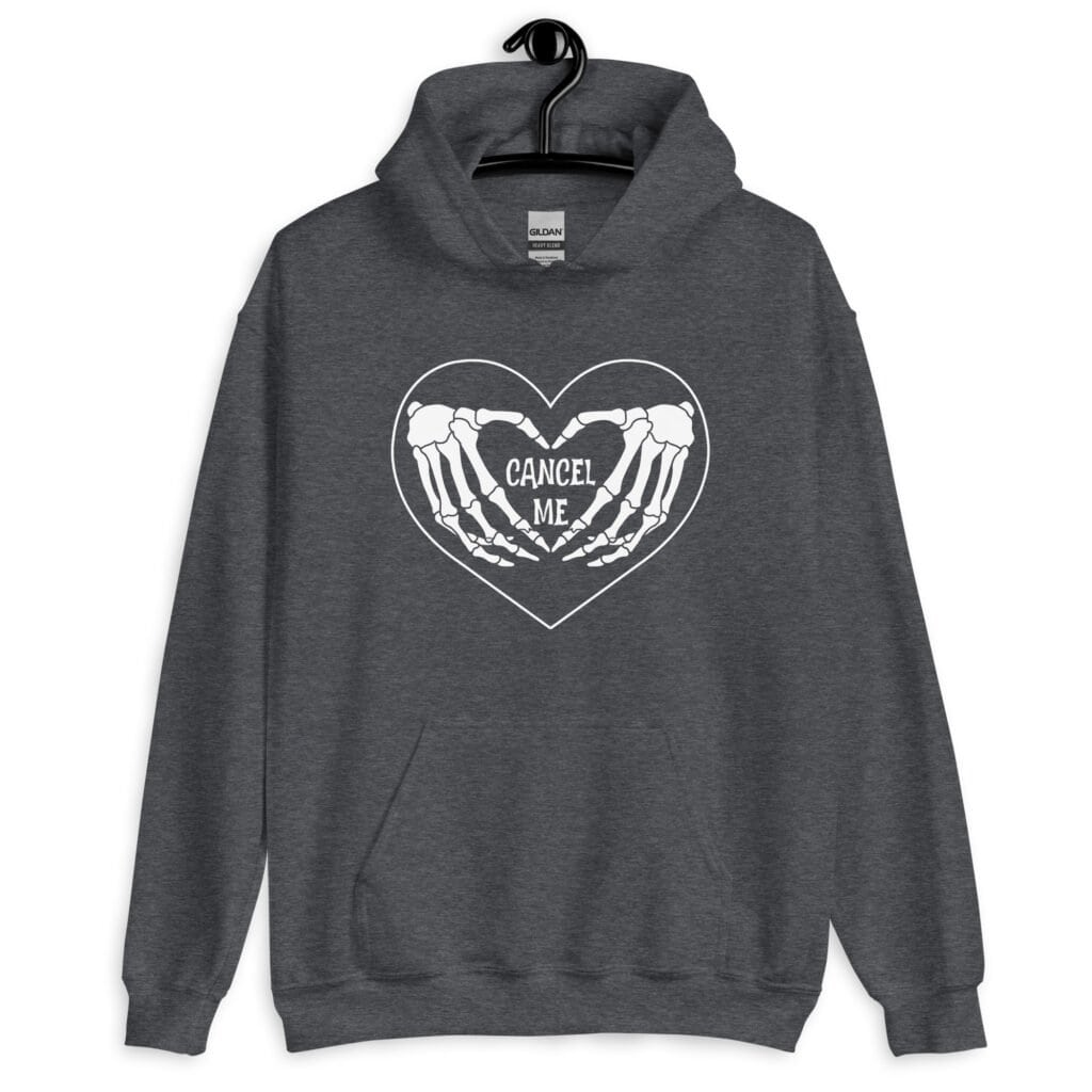 Dark heather grey hoodie sweatshirt with a heart and skeleton hands making a heart shape printed on the front. The words 'Cancel Me' are inside of the heart.