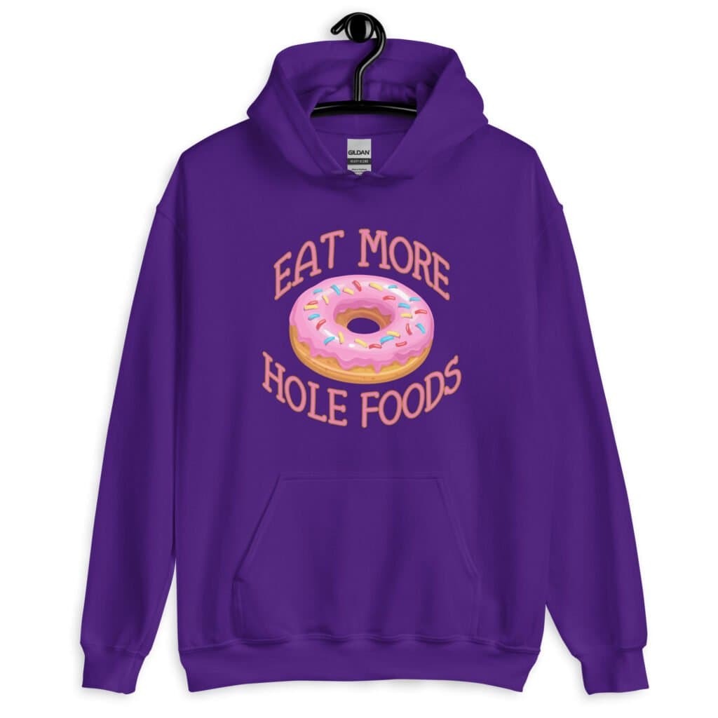 Purple hoodie sweatshirt with an image of a donut with pink icing and sprinkles and the text 'Eat more hole foods' printed on the front.