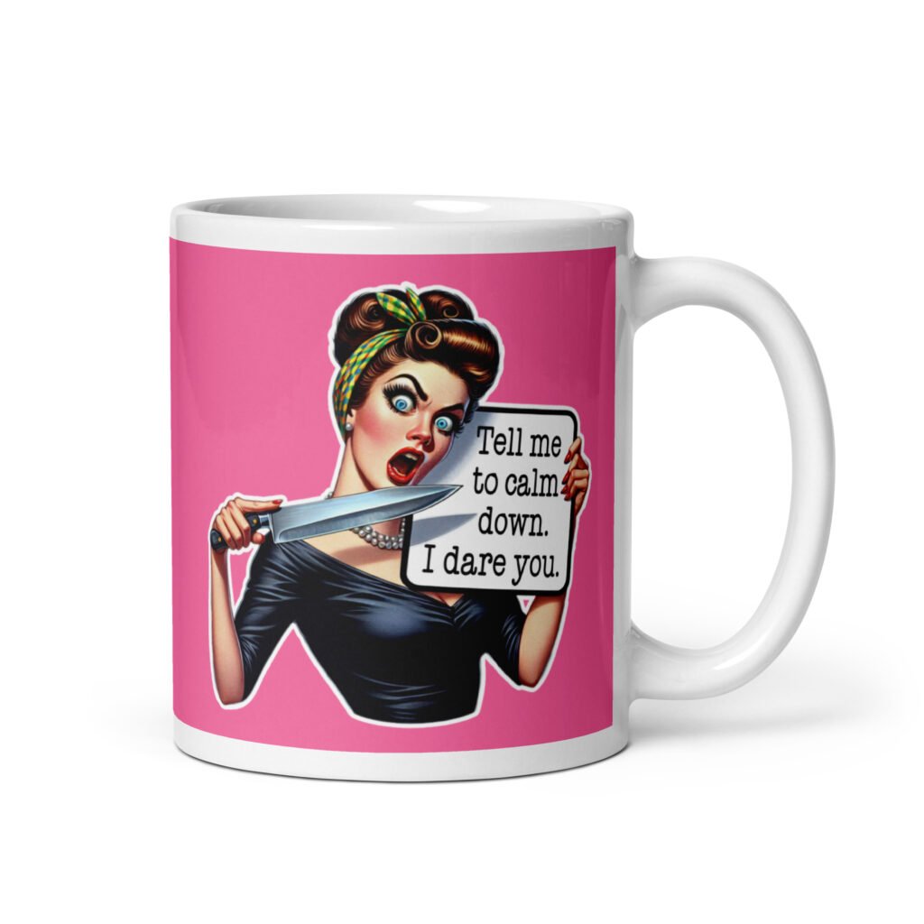 White ceramic coffee mug with pink color wrapped around. The mug has a graphic of an angry looking retro woman holding a knife and a sign. The sign says 'Tell me to calm down. I dare you.' The graphic is printed on both sides of the mug.