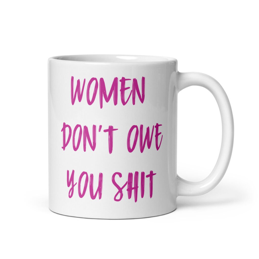 White ceramic coffee mug with the words 'Women don't owe you shit' printed on both sides in pink.