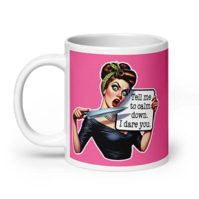 White ceramic coffee mug with pink color wrapped around. The mug has a graphic of an angry looking retro woman holding a knife and a sign. The sign says 'Tell me to calm down. I dare you.' The graphic is printed on both sides of the mug.