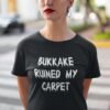Woman wearing a black t-shirt with the text 'Bukkake ruined my carpet' in drippy font printed on the front.