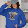 Woman wearing a royal blue hoodie sweatshirt with an image of a flower and the words Be kind above the flower in yellow bold block font. The words Of a bitch are smaller in script font under the flower. The design is printed on the front.