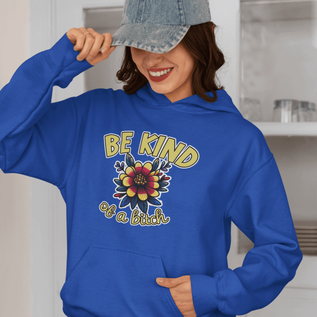 Woman wearing a royal blue hoodie sweatshirt with an image of a flower and the words Be kind above the flower in yellow bold block font. The words Of a bitch are smaller in script font under the flower. The design is printed on the front.