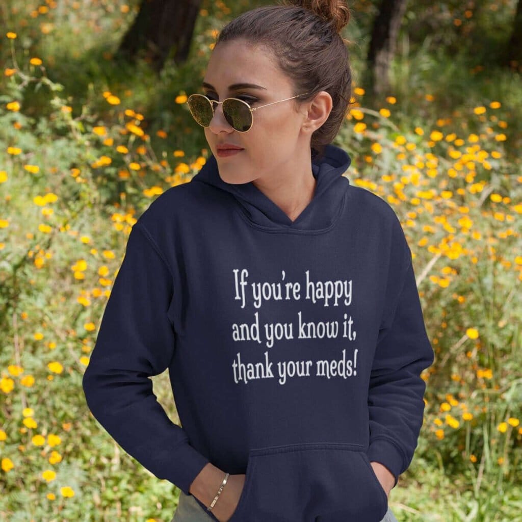 Woman standing near flowers wearing a navy blue hoodie sweatshirt with the text 'If you're happy and you know it, thank your meds!' printed on the front.