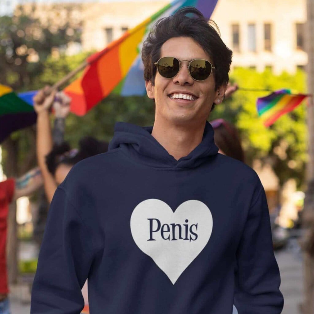 Man at a pride event wearing a navy blue hoodie sweatshirt with a heart image printed on the front. The word 'Penis' is inside the heart.