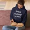 Woman reading a book while wearing a navy blue hoodie sweatshirt with the text 'Reading can seriously damage your ignorance.' printed on the front.