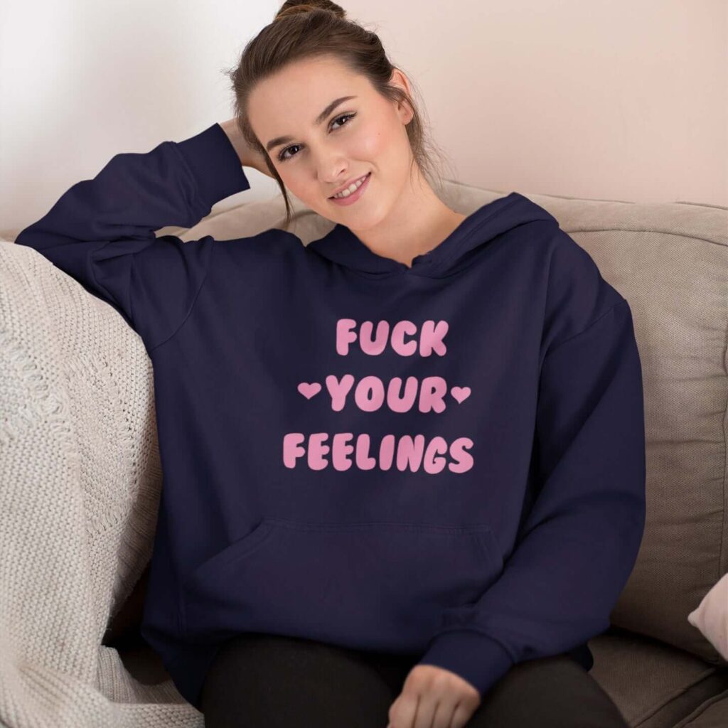 Woman wearing a navy blue hoodie sweatshirt with the text 'Fuck your feelings' printed on the front in pink. There are cute pink hearts next to the words.