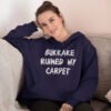 Woman wearing a navy blue hoodie sweatshirt with the text 'Bukkake ruined my carpet' in drippy font printed on the front.
