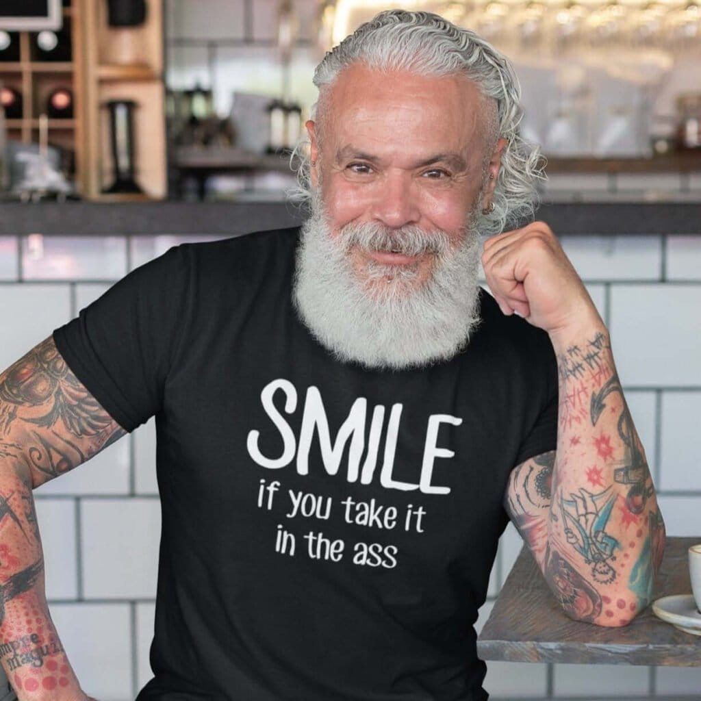 Handsome older man wearing a black t-shirt the text 'Smile if you take it in the ass' printed on the front. The word 'smile' is large and the words 'take it in the ass' are much smaller.