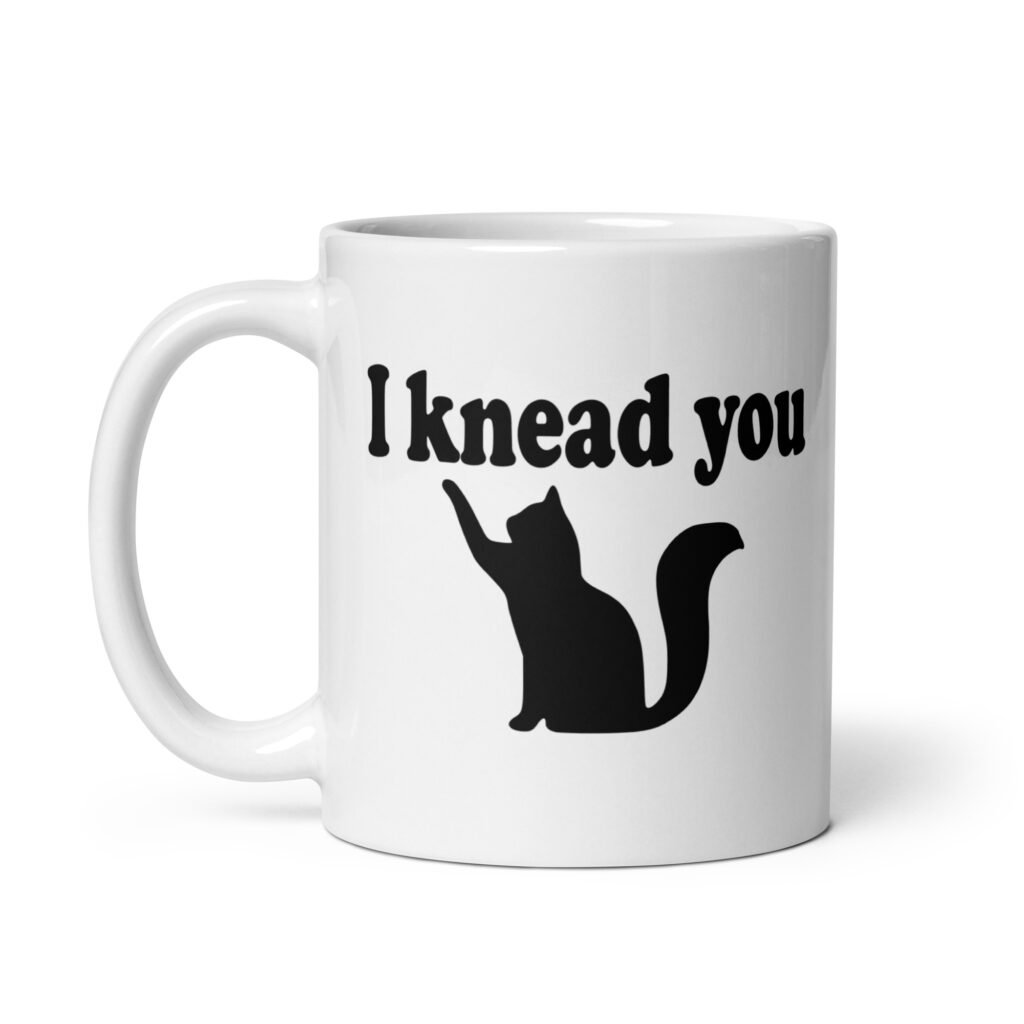 White ceramic coffee mug that has an image of a silhouette of a cat and the text 'I knead you' printed on both sides of the mug.
