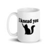 White ceramic coffee mug that has an image of a silhouette of a cat and the text 'I knead you' printed on both sides of the mug.