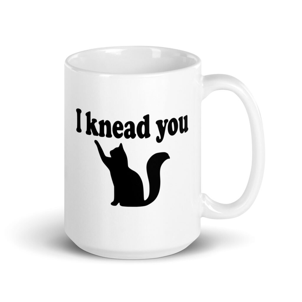 White ceramic coffee mug that has an image of a silhouette of a cat and the text 'I knead you' printed on both sides of the mug.