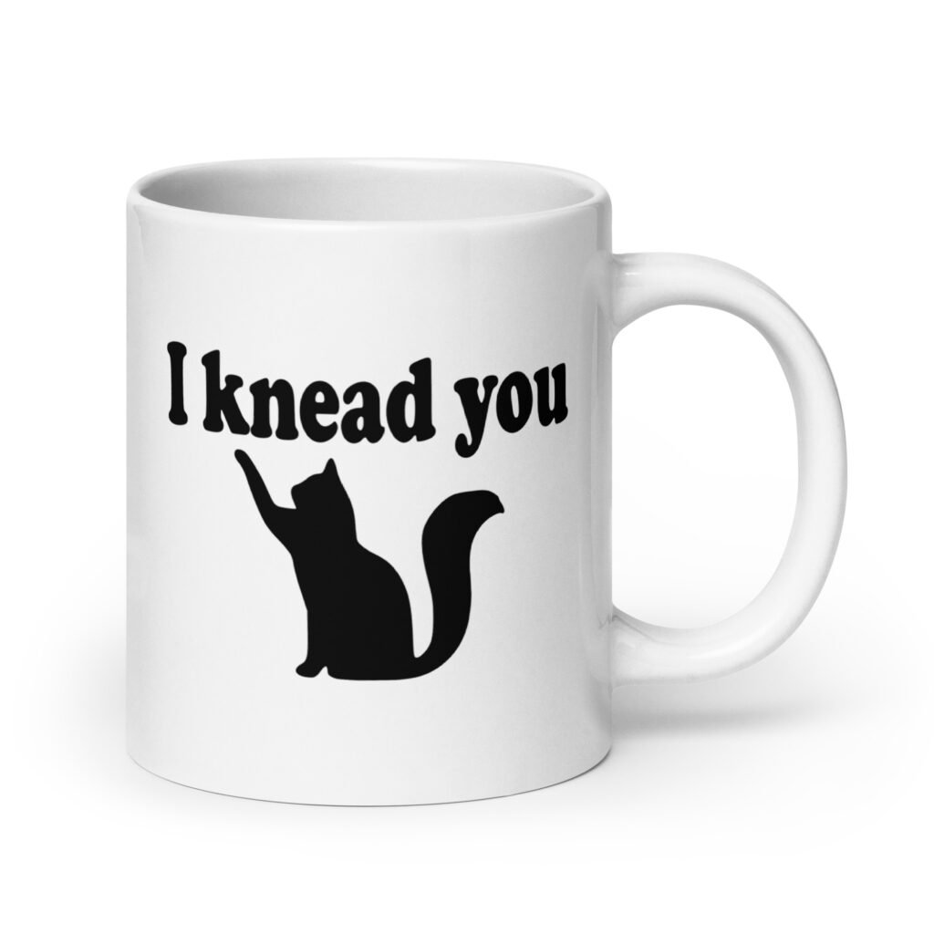 White ceramic coffee mug that has an image of a silhouette of a cat and the text 'I knead you' printed on both sides of the mug.