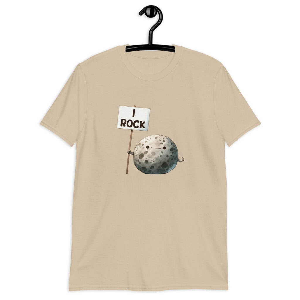 Sand color t-shirt that has an image of a grey rock that is holding a sign. The sign says 'I Rock.' The image is printed on the front of the shirt.