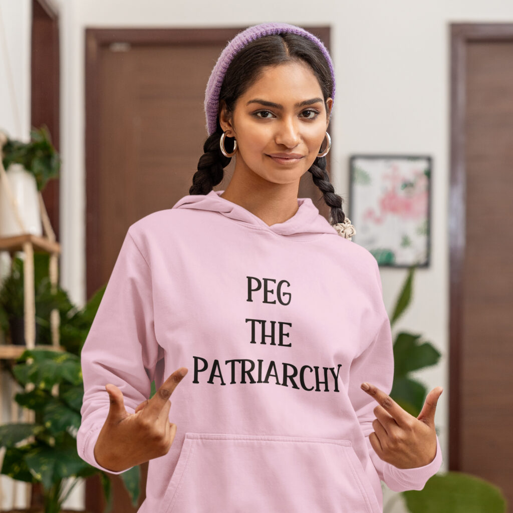 Woman wearing a light pink hoodie sweatshirt with the text 'PEG THE PATRIARCHY' printed on the front.