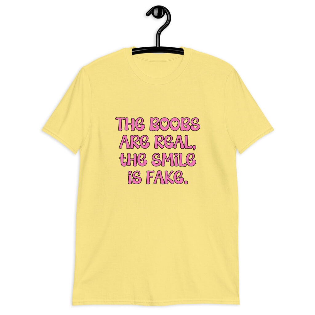 Cornsilk yellow t-shirt with the text 'The boobs are real, the smile is fake.' printed in pink on the front.