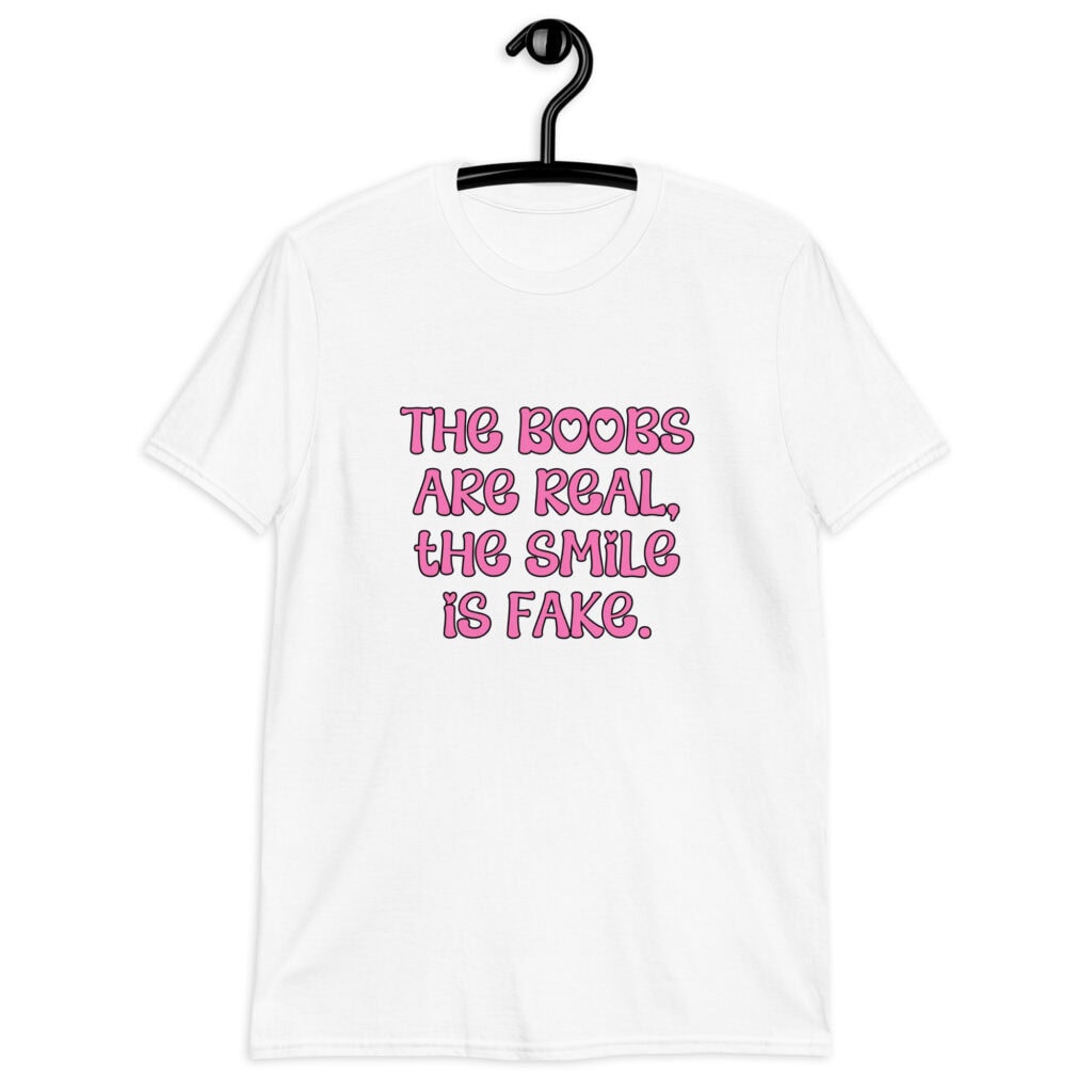 White t-shirt with the text 'The boobs are real, the smile is fake.' printed in pink on the front.