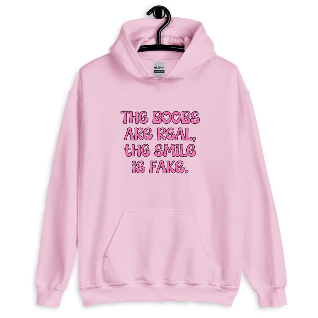Light pink hoodie sweatshirt with the text 'The boobs are real, the smile is fake.' printed in pink on the front.