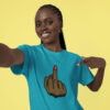 Smiling woman wearing a bright turquoise T‑shirt with a graphic of a brown hand showing the middle finger, red nail polish, against a yellow background.